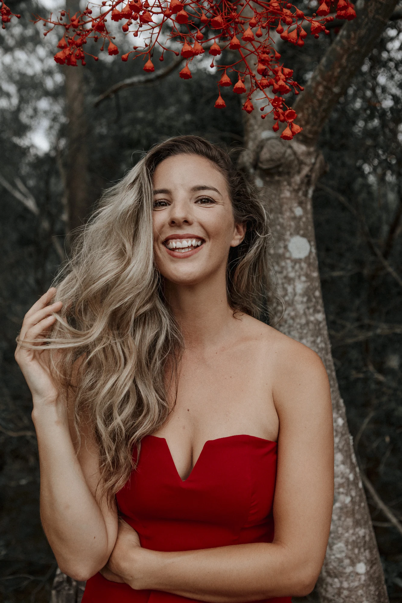 Yasmina smiling in a red dress beneath red flowering branches.